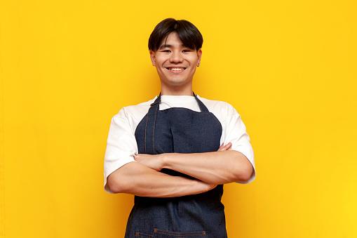 young asian guy barista in apron stands with arms crossed on yellow isolated background, korean male waiter in uniform looking at camera, service worker