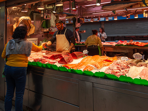 Málaga, Spain - 3rd November 2023: A customer paying for her purchase from a fresh fish stall in Mercado Central de Atarazanas, the food market in central Málaga. Another vendor is in the photo as are two potential customers. The stall sells wet fish such as Cod, Red Tuna (Atlantic Bluefish tuna) and Monkfish heads.