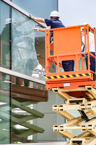 High-altitude work. Two blue-collar workers are using a lifting platform to repair broken glass on the office building's facade.