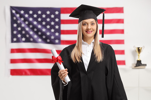 Female graduate student with a diploma wearing a graduation hat with USA flag in the background