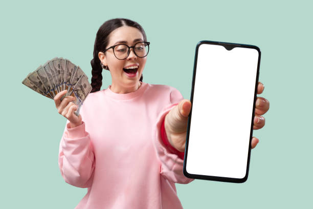 торговля на бирже. молодая кавказская выходная и радостная женщина в очках показывает веер денег и держит в руках смартфон с макетом. понят� - махать моделью стоковые фото и изображения