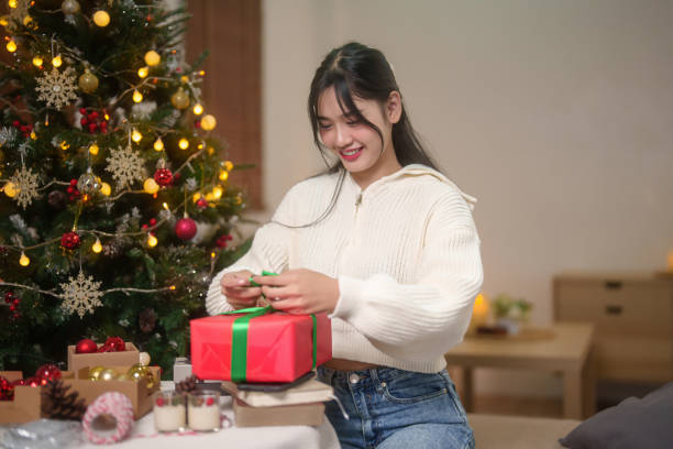 Happiness asian women in sweater is wrapping gift box and preparing toys ornament with bauble for decorating on christmas tree in living room while celebrate xmas tradition and holiday season at home. las mujeres asiáticas de la felicidad en el suéter están envolviendo la caja de regalo y preparando el adorno de los juguetes con adornos para decorar en el árbol de navidad en la sala de estar mientras celebran la tradición de la navidad y la tempora
