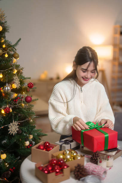 Happiness asian women in sweater is wrapping gift box and preparing toys ornament with bauble for decorating on christmas tree in living room while celebrate xmas tradition and holiday season at home. las mujeres asiáticas de la felicidad en el suéter están envolviendo la caja de regalo y preparando el adorno de los juguetes con adornos para decorar en el árbol de navidad en la sala de estar mientras celebran la tradición de la navidad y la tempora