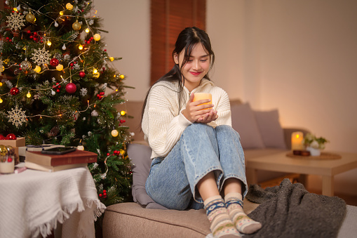 Happiness asian women in sweater sitting on couch near the christmas tree in living room and holding candles for decoration to preparing for celebrate xmas tradition and holiday season at home.