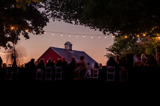Long exposure of an outdoor wedding ceremony at twilight and guests having a fun celebrating. Motion blur makes any people unrecognizable. Barn and string lights set the scene for the style.