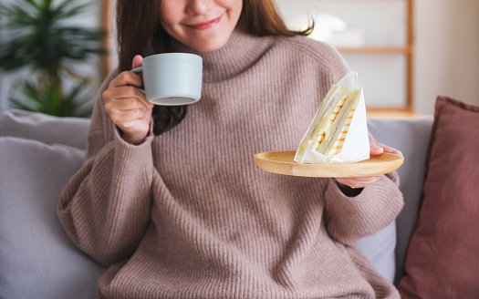 Closeup image of a woman holding coffee cup and sandwich at home
