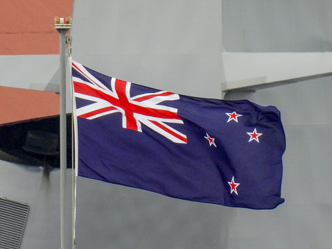 The flag of New Zealand flies on the bow of HMNZS Aotearoa docked at Garden Island in Sydney Harbour.  She is a Polar Class Logistics Support ship of the Royal New Zealand Navy.  In the background is HMAS Warramunga, an Anzac Class frigate of the Royal Australian Navy.  This image was taken on an overcast and windy afternoon on 25 November 2023.