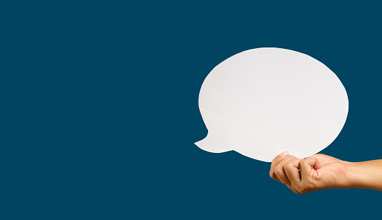 Close-up of hand holding a blank white speech bubble while standing against a blue background. Space for text