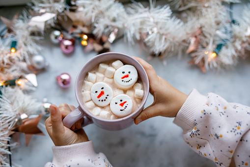 Unrecognizable little child girl holding cup with hot chocolate with marshmallows as snowman. Kid sitting near Christmas decorated window with lights