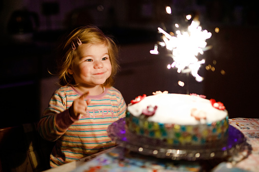 Adorable little toddler girl celebrating second birthday. Baby child eating marshmellows decoration on homemade cake, indoor. Happy healthy toddler is suprised about firework sparkler on cake.