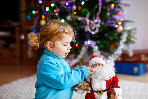 Adorable toddler girl playing with gifts and Christmas Santa Claus toys. Little child having fun with decorated and illuminated Xmas tree with lights on background. Happy healthy funny baby girl