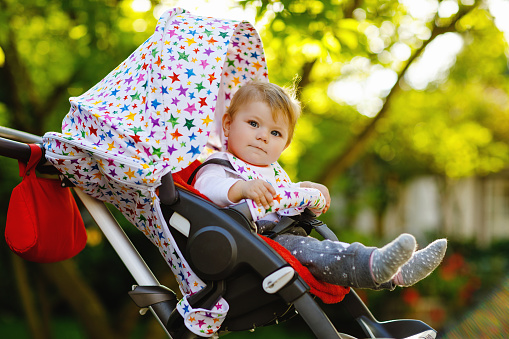 Cute healthy little beautiful baby girl sitting in the pram or stroller and waiting for mom. Happy smiling child with blue eyes. With green tree background. Baby daughter going for a walk with family.