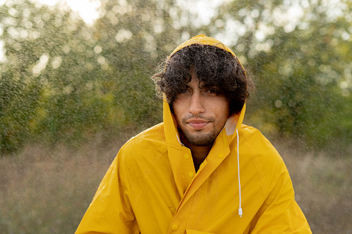 Sad man freezes in the rain wearing a yellow raincoat in the park. Handsome male under rain outdoors. Guy has a tired expression during the rainy weather. Copy space