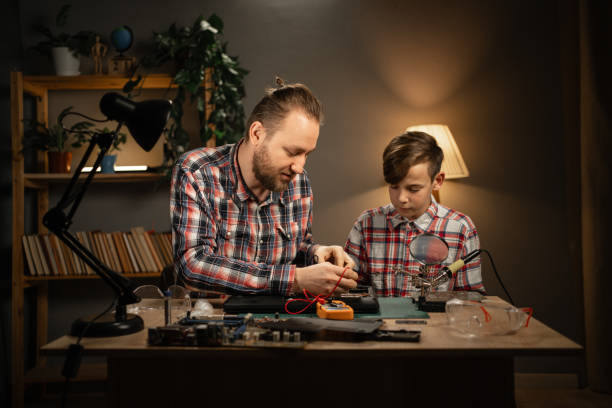 istock Father and his son using soldering iron and multimeter for fixing motherboard from laptop. Two men sitting together at table and repairing computer at home. 1814208470
