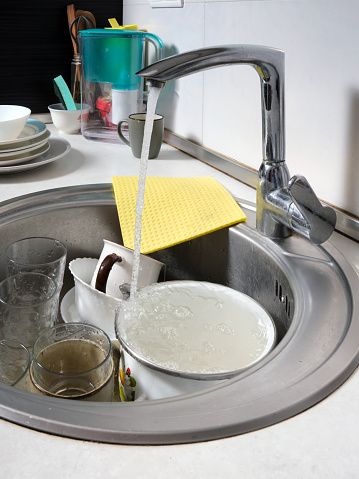 Close-up of a kitchen sink with dirty dishes. Water flows from an open tap. Reluctance to do routine housework. Vertical image.