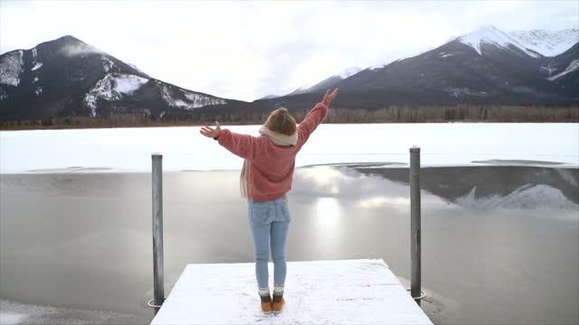 Slow Motion: Young woman standing arms outstretched near frozen mountain relaxing in winter vacations