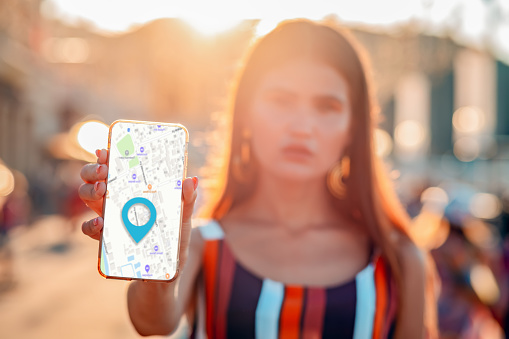 A blurred woman holds a cellphone with an online map app, hand close-up. In the background is a city street in sunset light. The concept of online navigation and modern technologies.