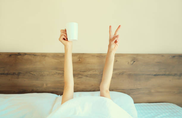 istock Cheerful lazy woman waking up after sleeping lying in soft comfortable bed showing empty cup coffee stretching her hands up from under the blanket in bedroom at home 1814422343