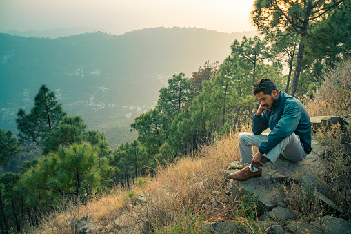A young man of Indian descent sits on a rock and contemplates deeply in the tranquil mountain of Himachal Pradesh, India.