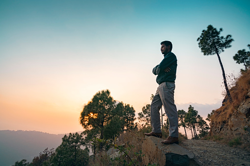 An Asian/Indian young man looks at the sunset view standing on a rock in the tranquil mountain of Himachal Pradesh, India.