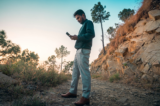 A young man of Indian descent uses his phone while standing on a dirt road in the mountains.