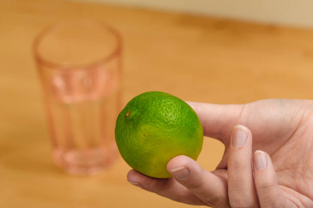 uma cal verde segurada na mão, um copo de água ao fundo - close up women horizontal citrus fruit - fotografias e filmes do acervo