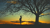 Side view of silhouette pregnant woman standing with hands on stomach by single bare tree against moody sky during sunset