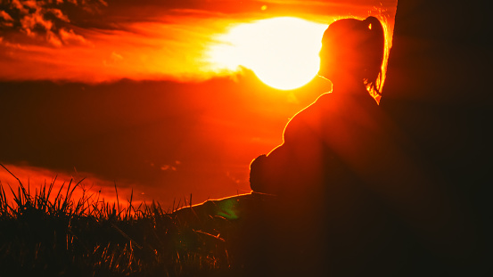 Tranquil silhouette,A relaxed pregnant woman leaning against a tree trunk,bathed in the warm glow of bright orange sunlight during a picturesque sunset