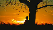 Side view of silhouette pregnant woman with hands on stomach leaning on single bare tree against dramatic sky during sunset