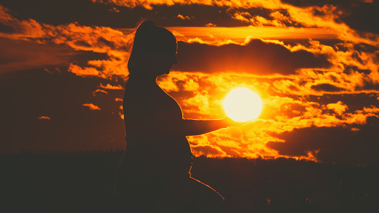 In a captivating profile view silhouette,a pregnant woman holds the sun against a dramatic sky during sunset. This emotive scene beautifully captures the serene beauty of expectancy and the tranquil connection with nature