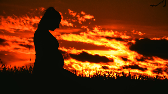 In a poignant silhouette,a pregnant woman kneels with hands on her stomach against a dramatic sky during sunset. This emotive scene captures the serene beauty of expectancy and the tranquil connection with nature in the fading light