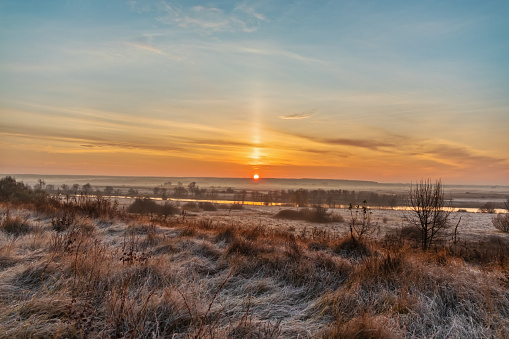 Scenic view of the rising Sun over a river valley with trees and meadows on a foggy morning. Landscape of late autumn or early winter with frost on the grass. Light pillar - an atmospheric optical phenomenon, a vertical beam of light from the Sun