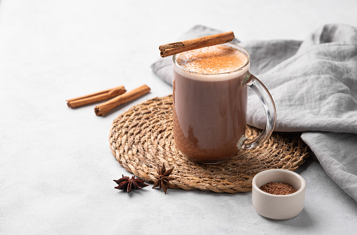 Glass mug with hot chocolate and milk foam on a light background with cinnamon sticks, anise star and cocoa powder. Warming winter  drink concept. Free space for text.