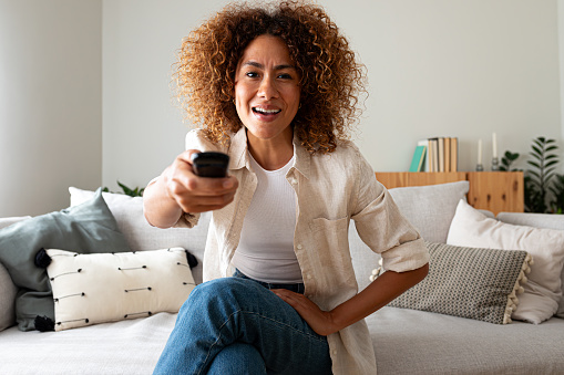 Happy African American woman watching TV sitting on the sofa using remote control to change channel looking at camera with surprise expression. Lifestyle concept.