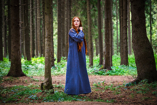 Red haired young woman in a linen blue dress is escape in the middle of the forest in the rays of the sun. The girl experiences emotions of despair left alone in the forest.