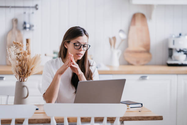 young female tutor in glasses, sitting at a desk at home, working on a laptop, making a video call, talking to  student, checking her homework.  Pretty American girl learning remotely during pandemic