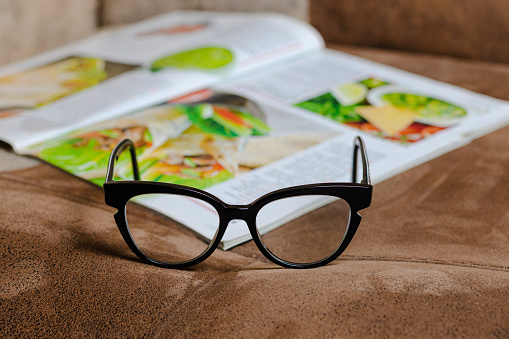 Women's glasses in a black frame on the sofa, against the background of a magazine with recipes
