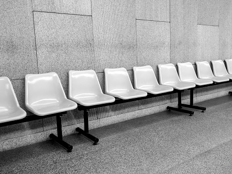 Black and white photo scene of row of empty waiting chair with white plastic seats with black iron legs seats on grey tiles and concrete wall background inside the modern building with nobody.