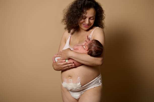 autentyczna mama w bieliźnie, uśmiecha się, patrząc na swoje nowo narodzone dziecko, które trzyma, pokazuje swoje ciało z bliznami poporodowymi, wadami - cesarka zdjęcia i obrazy z banku zdjęć