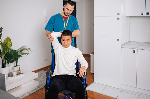 A young Caucasian man is captured helping a mid-adult Chinese man in a wheelchair with arm stretches, facilitating physical therapy in a caring home environment