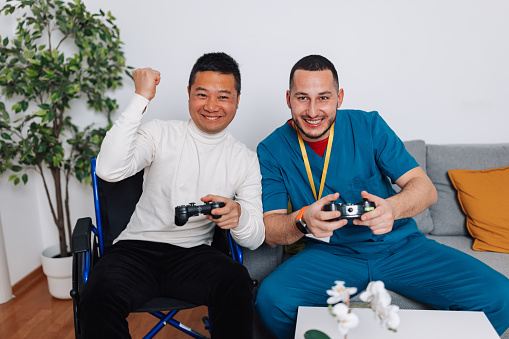 A photo shows a young Caucasian man and a mid-adult Chinese man, wheelchair-bound, engaged in a lively video game battle, each wielding a joystick with concentration and jo