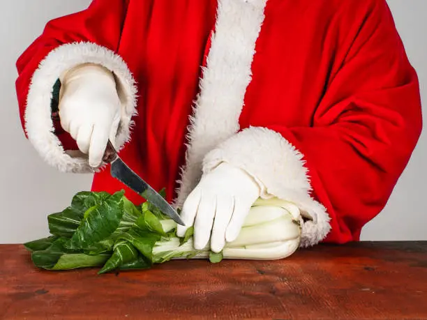 Santa Claus cuts fresh bok choy with a knife on a wooden table. Healthy eating of vegetables in winter.