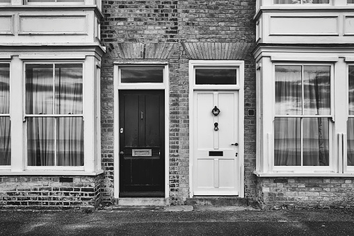 Matt monochrome view of a pair of wooden from doors seen on brick built terraced houses overlooking a seaside position in the UK. The seaside reflection can just be seen in the bay windows.