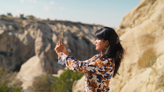 A multiracial beautiful female tourist is using her smart mobile phone to take photos and videos of nature and rock formations in Cappadocia in Türkiye Turkey.