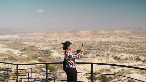 A multiracial beautiful female tourist is using her smart mobile phone to take photos and videos of nature and rock formations in Cappadocia in Türkiye Turkey.