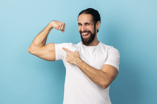 Man bodybuilder with beard wearing white T-shirt pointing finger at his arm biceps, proud and satisfied with muscular build, leadership skills. Indoor studio shot isolated on blue background.