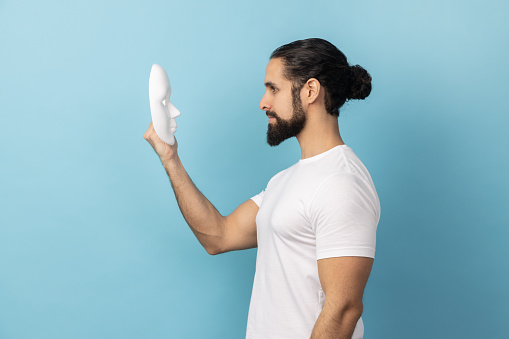 Side view of man with beard wearing white T-shirt holding and looking at white mask with attentive look, trying to understand hiding personality. Indoor studio shot isolated on blue background.