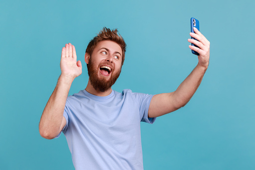 Portrait of positive optimistic bearded man talking on video call and waving hello gesture, having online conversation on mobile phone. Indoor studio shot isolated on blue background.