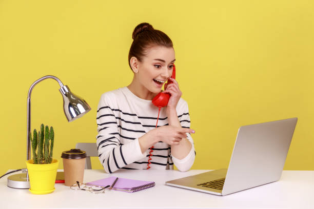 istock Woman making call on landline phone working on laptop sitting at office pointing at notebook display 1814901253