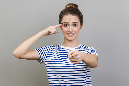You are idiot. Woman wearing striped T-shirt showing stupid gesture and pointing to camera, blaming for insane plan, crazy idea, dumb suggestion. Indoor studio shot isolated on gray background.